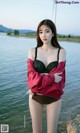 A woman in a red jacket and black bra posing by the water.