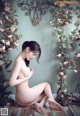 A naked woman sitting on the floor in front of a wall of flowers.