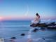 A woman sitting on a rock in the ocean at sunset.